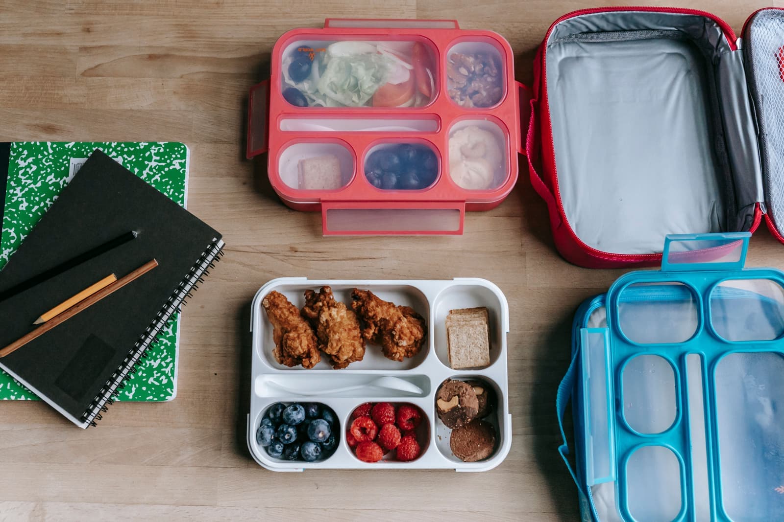 Packed lunch boxes beside notebooks and pencils on a wooden table, seen from above